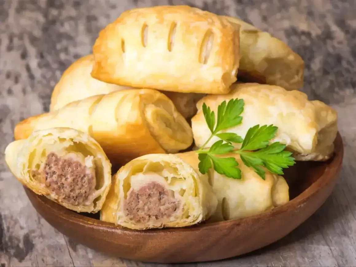 Stos pieczonego ciasta francuskiego z kiełbasą w środku, ozdobiony gałązką pietruszki.