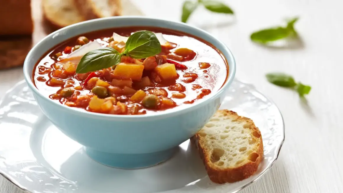 Aksamitna zupa solferino w błękitnej misce, udekorowana bazylią i parmezanem, obok kromka chleba.