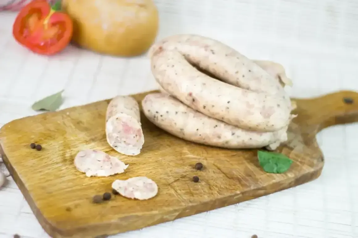 Biała kiełbasa na desce, gotowa do przygotowania według przepisu na 5 kg. W tle pomidor i cebula.