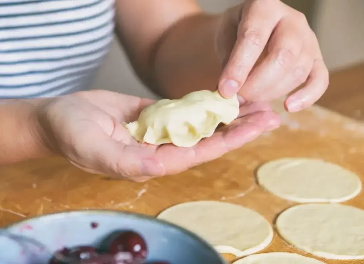 Dłonie formują pieroga z ciasta na pierogi bez jajka. Na blacie leżą okrągłe placki z ciasta i miska z wiśniami.