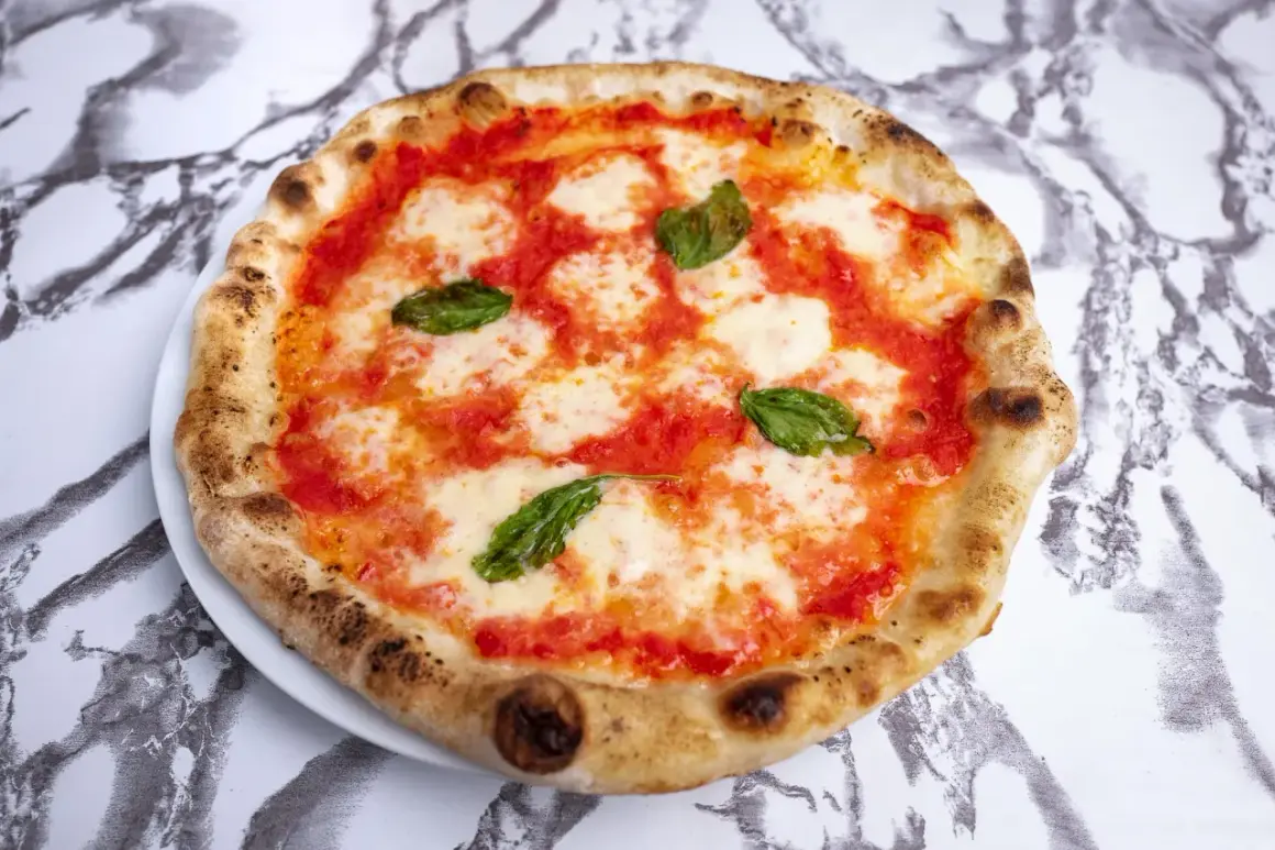 Pyszna pizza napoletana z pomidorami, mozzarellą i bazylią na białym talerzu, na tle marmurowego wzoru.