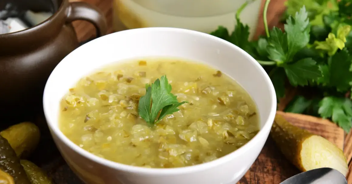 Zupa ogórkowa przepis babci. Aromatyczna, domowa zupa z kiszonych ogórków, podana w białej miseczce z gałązką natki pietruszki. Obok leżą ogórki kiszone.