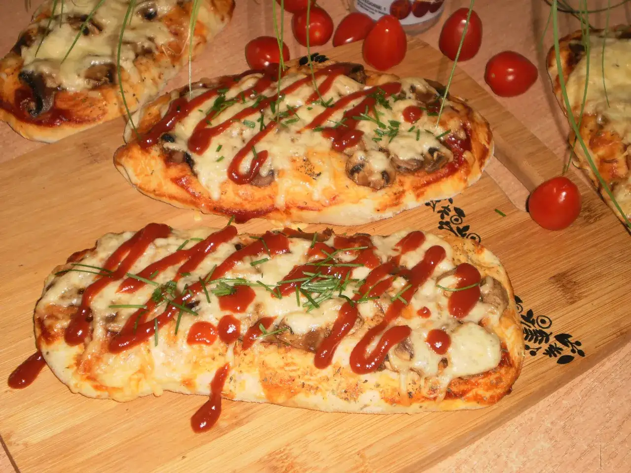 Dwa podłużne, zapieczone ciasta na pizzę z pieczarkami, serem i keczupem, ozdobione szczypiorkiem.