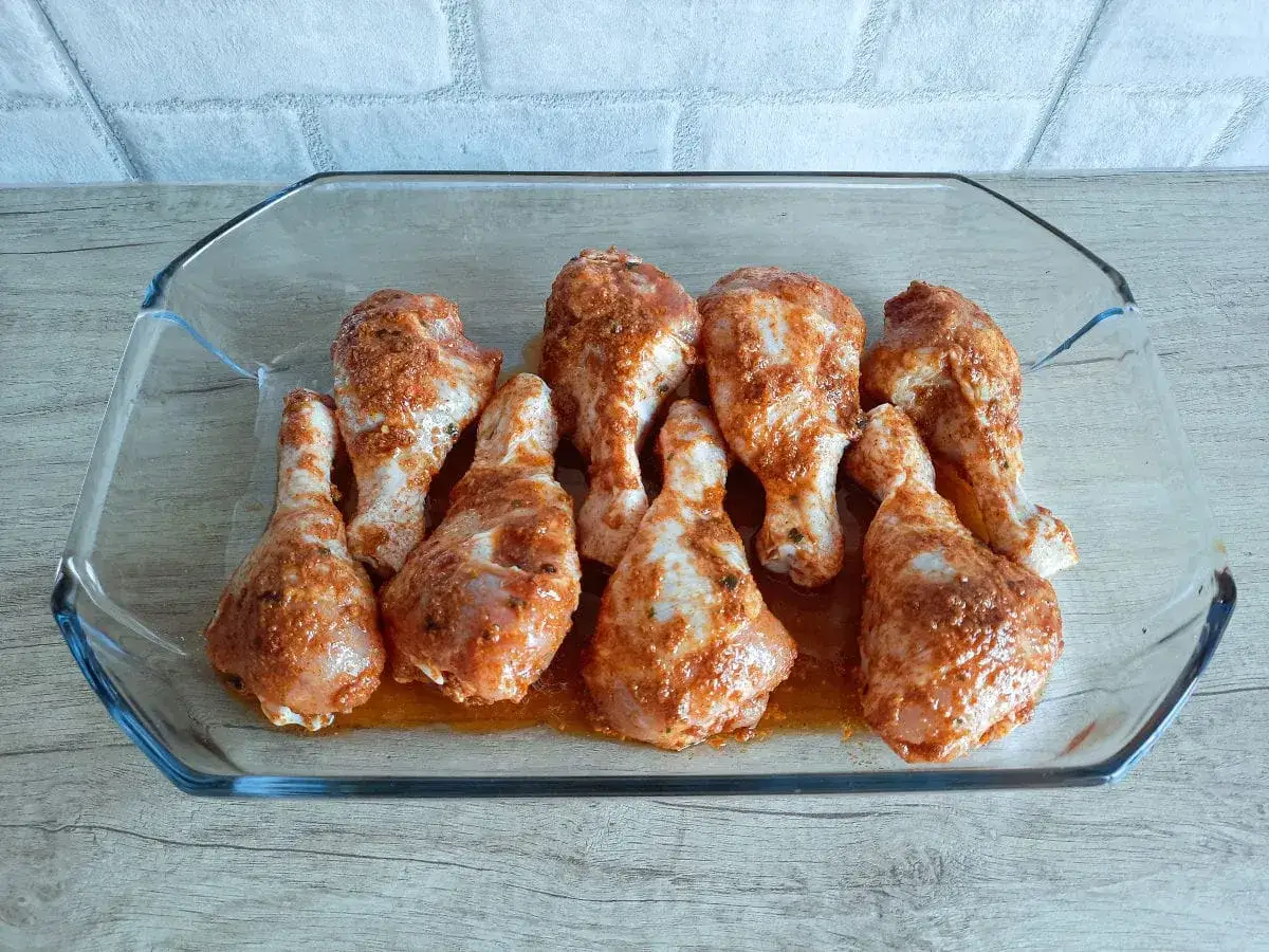 Gotowe do pieczenia pałki z kurczaka w przyprawach, ułożone w szklanym naczyniu.