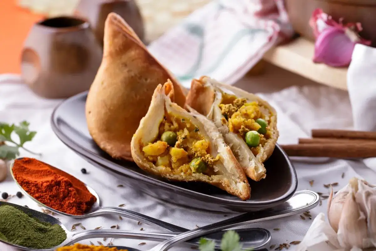 Chrupiący indyjski pierożek samosa, przekrojony na pół, ukazuje bogate nadzienie z ziemniaków i groszku. Obok przyprawy: papryka, curry, czosnek.