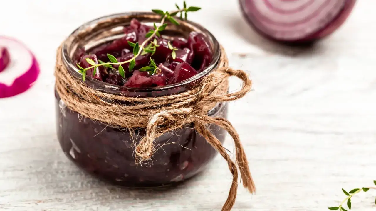 Słoik z domowym chutney z czerwonej cebuli, ozdobiony sznurkiem i gałązką tymianku. Obok widać plasterki czerwonej cebuli.
