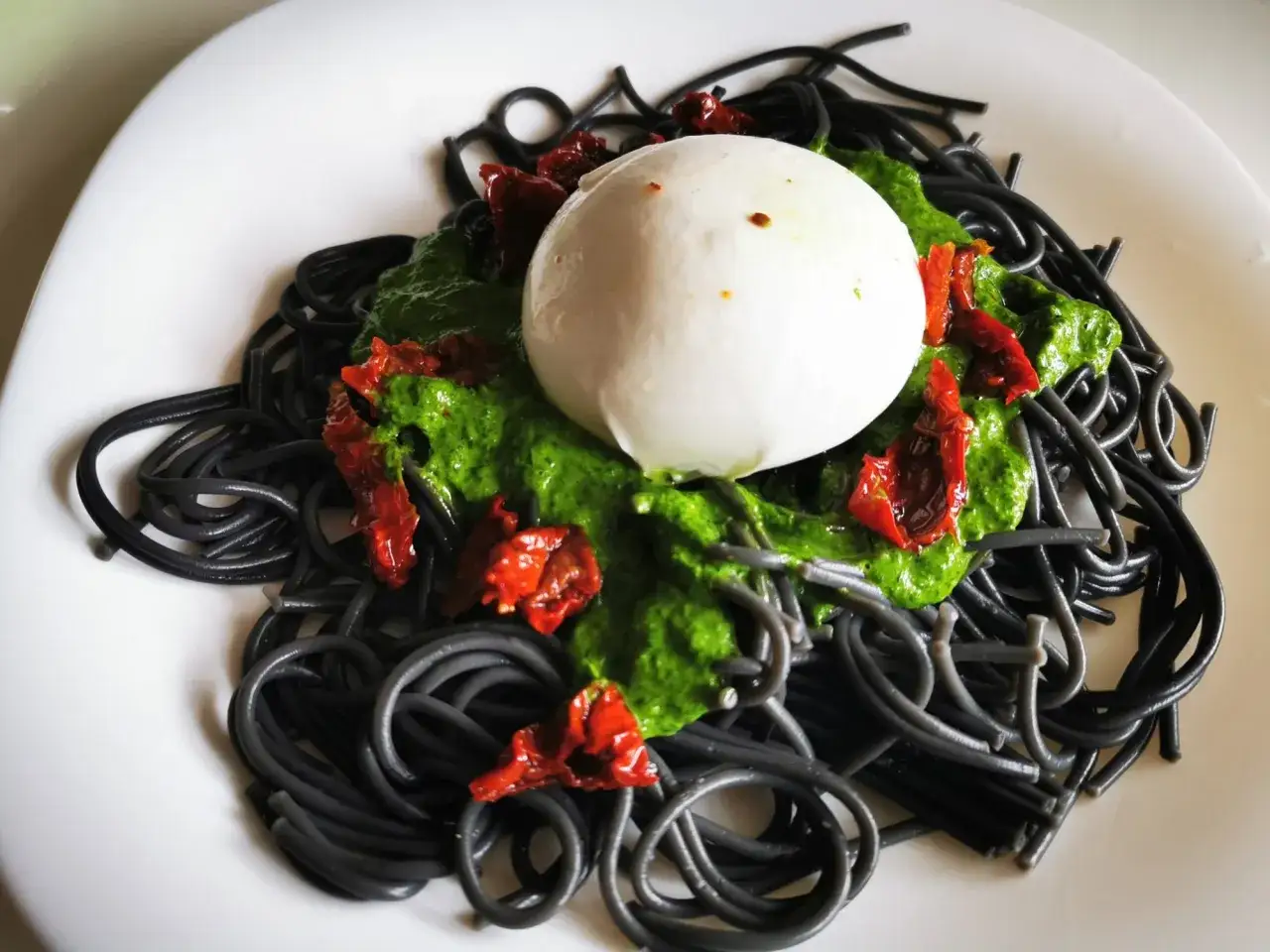 Czarny makaron z zielonym pesto, suszonymi pomidorami i kulką mozzarelli.