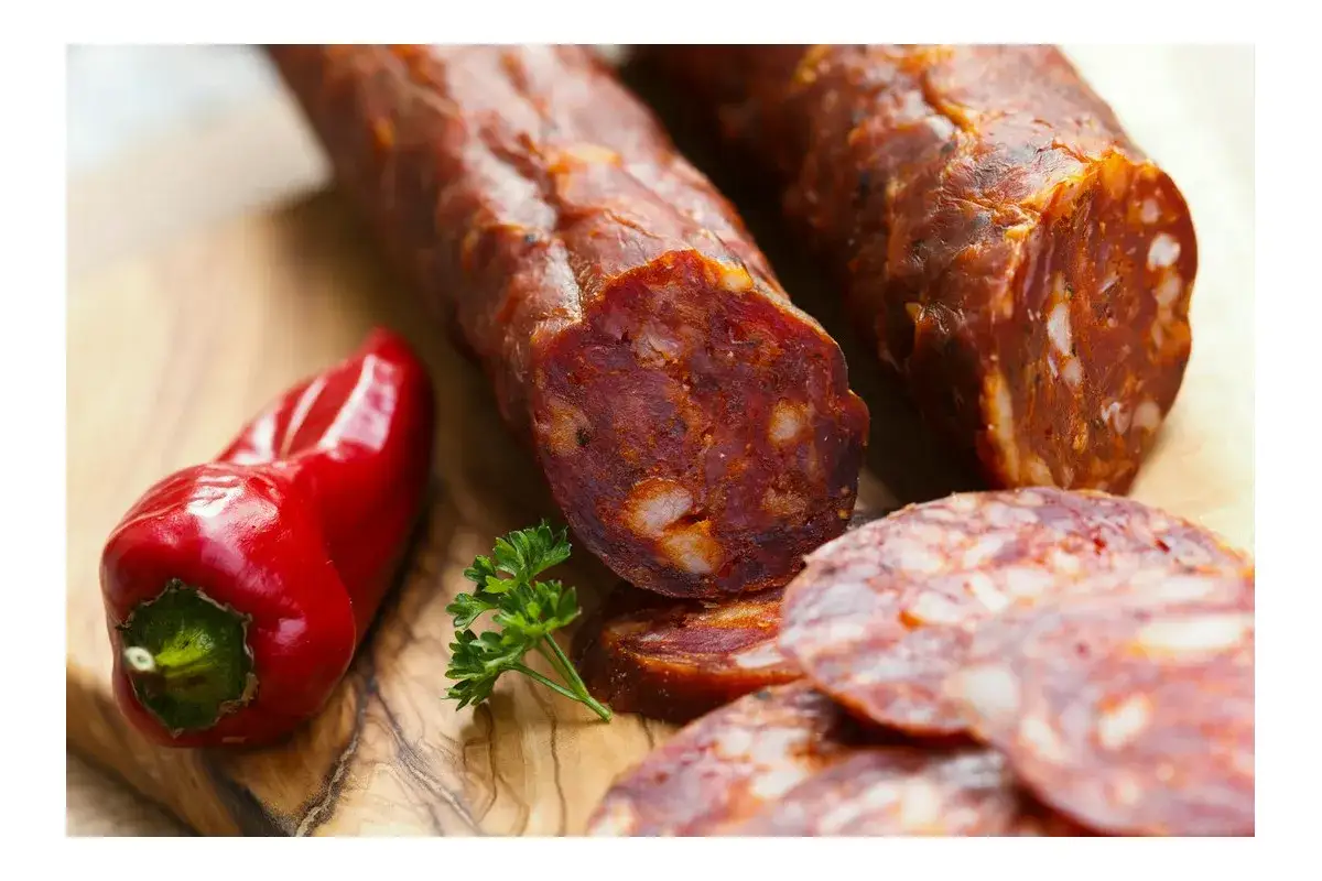 Kiełbasa chorizo w plastrach i w całości, obok czerwonej papryczki i natki pietruszki na desce.