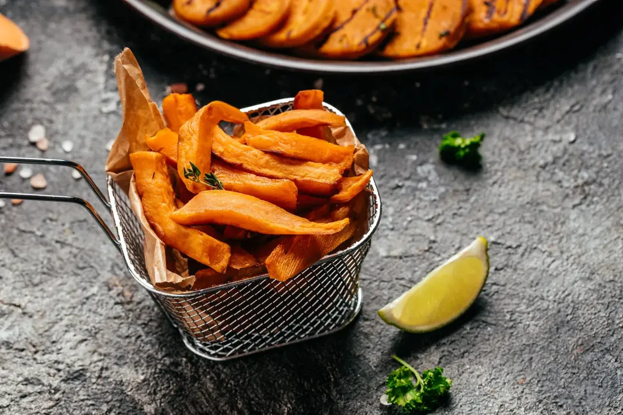 Chrupiące bataty z airfryer w metalowym koszyczku, ozdobione gałązką tymianku, obok ćwiartki limonki i natki pietruszki.