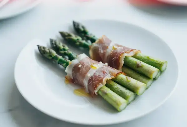 Zielone szparagi owinięte w cienkie plastry szynki parmeńskiej, ułożone na białym talerzu.