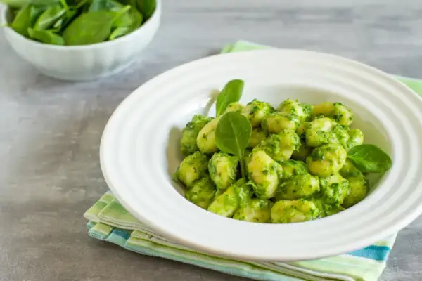 Puszyste gnocchi z pesto, podane w białej misce, ozdobione świeżymi listkami szpinaku. Obok miseczka ze świeżym szpinakiem.