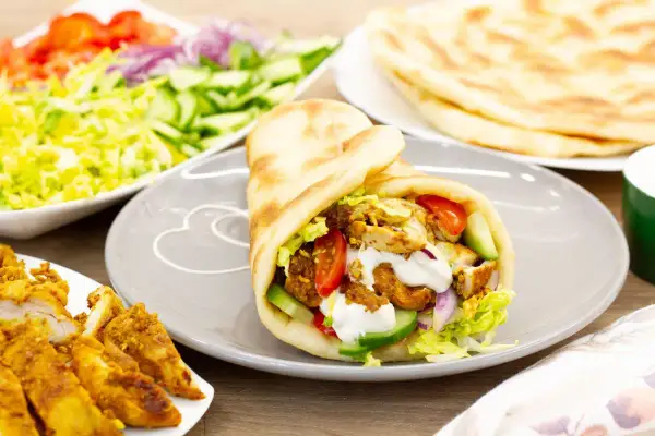 Pyszna shoarma drobiowa w picie z warzywami i sosem. Obok talerz z dodatkami: sałatka, pomidory, ogórki, cebula i placki chleba.