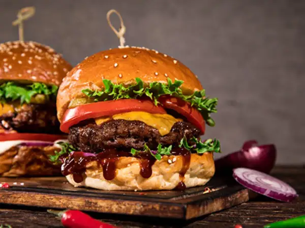 Dwa soczyste burgery z serem, sałatą i pomidorem na desce, obok cebuli i papryczki chili.