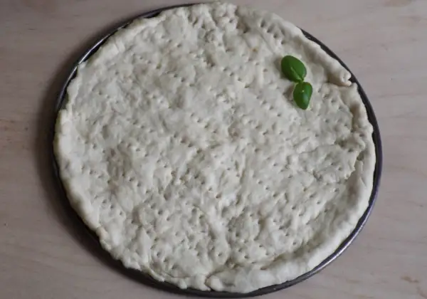 Surowy placek na pizzę w okrągłej formie, z kilkoma listkami bazylii. Proste ciasto na pizzę zawsze wychodzi.