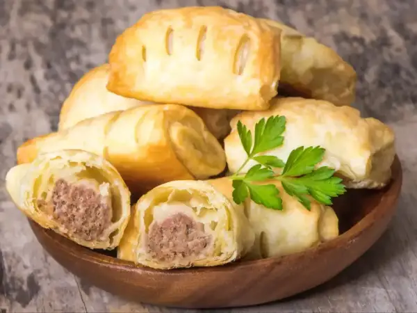 Stos pieczonego ciasta francuskiego z kiełbasą w środku, ozdobiony gałązką pietruszki.