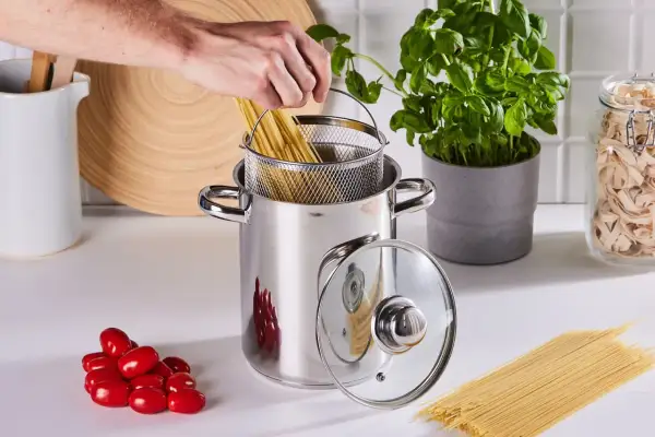 Ręka wkłada spaghetti do garnka, by ugotować je idealnie al dente. Obok pomidorki i świeża bazylia.