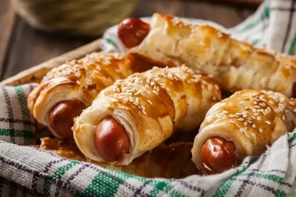 Chrupiące parówki w cieście francuskim z serem żółtym, posypane sezamem, ułożone w koszyku na kratkowanej ściereczce.