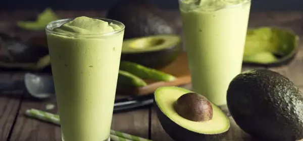 Dwa szklanki kremowego smoothie z awokado, idealna przekąska na upalne dni. Obok leżą świeże awokado i zielone słomki.