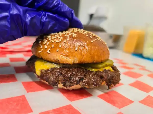 Rękawiczka w niebieskiej rękawiczce dociska pysznego smash burgera z serem i ogórkiem. Oto jak wygląda smash burger co to!