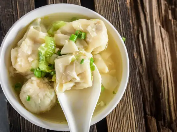Aromatyczna zupa wonton z zieloną cebulką i kawałkami selera, podana w białej miseczce na drewnianym blacie.