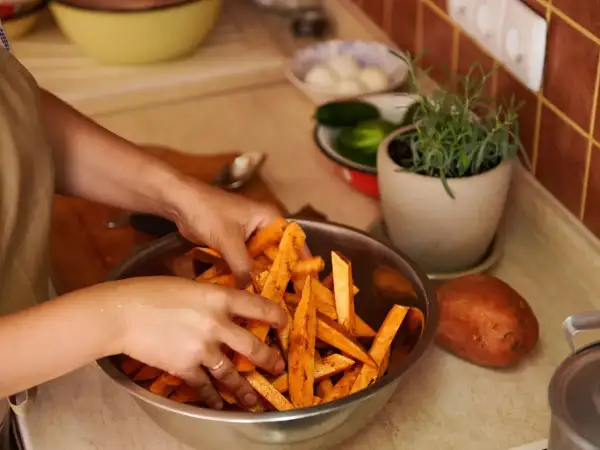 Ręce mieszają pokrojone bataty w misce, przygotowując je do pieczenia. Obok leży cały batat, a w tle zioła i warzywa, jakby czekały na sos belgijski.