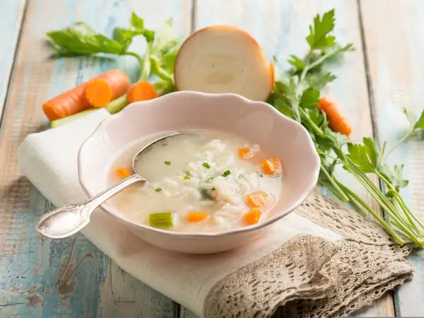 Aromaticzna zupa na rosole z ryżem, marchewką i selerem. Obok miseczki świeże warzywa: marchew, cebula, natka pietruszki.