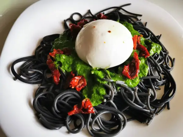 Czarny makaron z zielonym pesto, suszonymi pomidorami i kulką mozzarelli na białym talerzu.