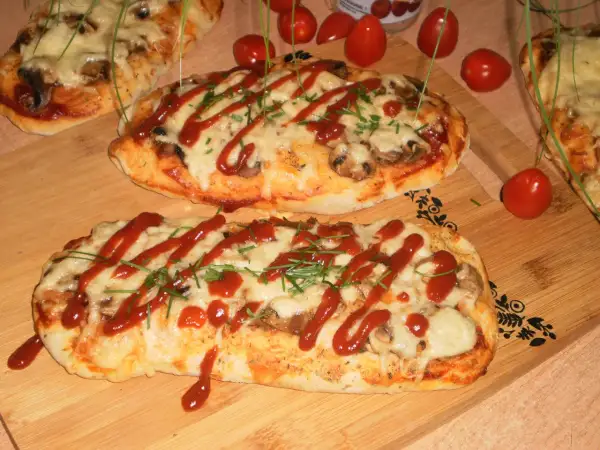 Dwa podłużne, zapieczone ciasta na pizzę z serem, pieczarkami i keczupem, ozdobione szczypiorkiem.