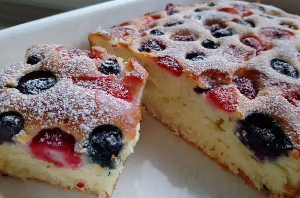 Puszyste ciasto na maślance z jagodami i malinami, posypane cukrem pudrem.
